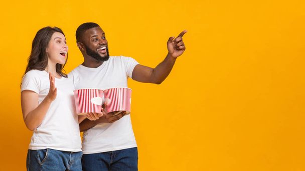 Excited interracial couple holding popcorn backets and pointing at copy space - Photo, Image