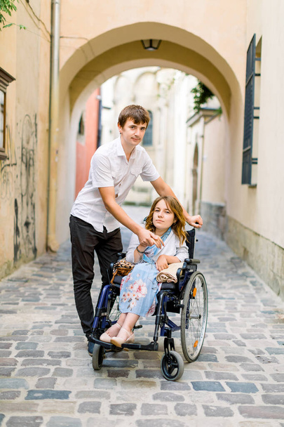 Young handsome smiling man and his pretty wife in wheelchair posing together on the background of old city center, arch of the ancient building. Love, disability concept - Photo, image