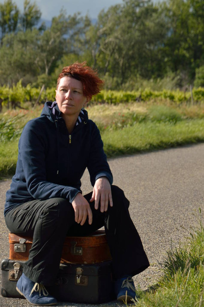 selfconfident red-haired woman on vacation sitting on her suitcases on a road and waiting - Photo, Image