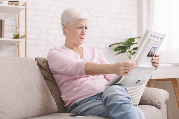 Vanhempi nainen siristelee ja pitelee sanomalehteä kaukana silmistä
 - Valokuva, kuva