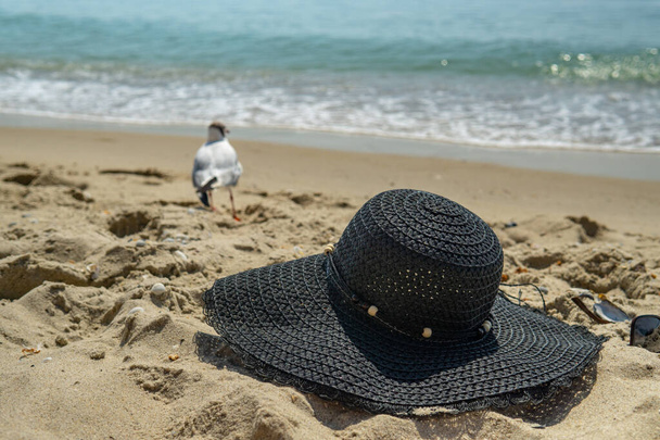 Letní prázdniny relaxovat na pláži abstraktní pozadí. Dámský klobouk na pláži písek, na pozadí moře a racků. - Fotografie, Obrázek