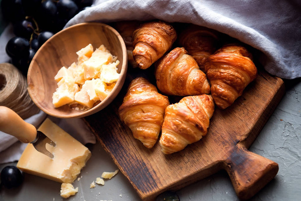 Eredeti ízletes francia croissant sajttal és szőlővel a fa asztalon. vajas pelyhes viennoiserie kenyér roll jellegzetes félhold alakú. Sajt tálban, kés, vágódeszka mint háttér. - Fotó, kép