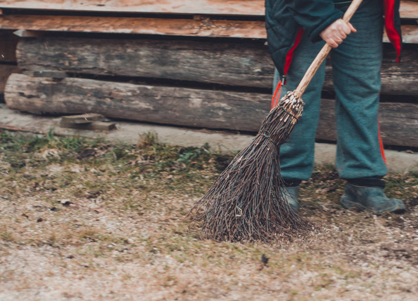 A woman in a village cleans a yard near her house with a birch broom. - 写真・画像