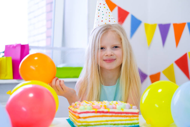 Blond blank meisje glimlachend naar de camera in de buurt van verjaardag regenboog taart. Feestelijke kleurrijke achtergrond met ballonnen - Foto, afbeelding