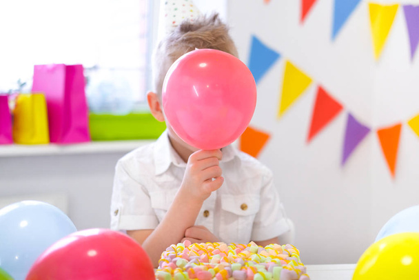  Blonde caucasian boy hid behind a red balloon near birthday rainbow cake. Festive colorful background. Funny birthday party - Photo, image