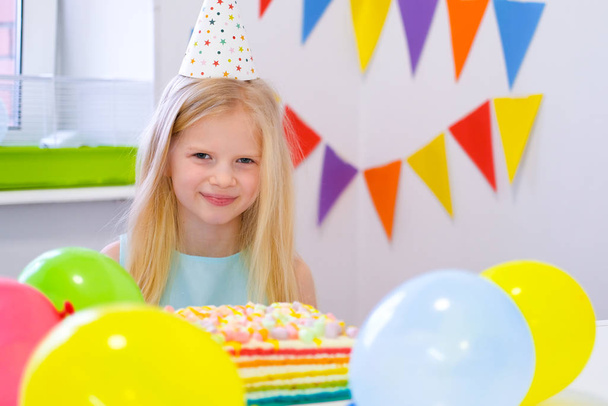 Blonde caucasian girl smiling at camera near birthday rainbow cake. Festive colorful background with balloons - 写真・画像