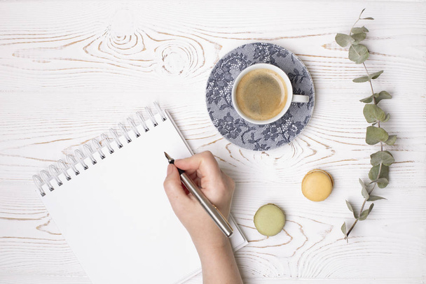 Ένα γυναικείο χέρι κρατά ένα στυλό πάνω από ένα καθαρό ημερολόγιο με ελατήριο, ένα φλιτζάνι καφέ και αμυγδαλωτά και ένα κλαδί ευκαλύπτου πάνω σε ένα λευκό άθλιο ξύλινο τραπέζι. Μόδα επίπεδη lay - Φωτογραφία, εικόνα