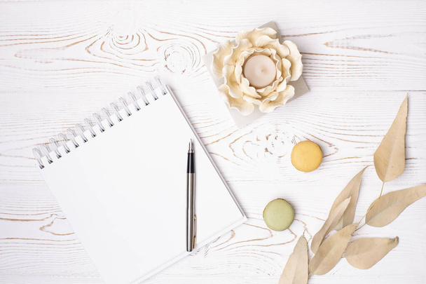 Top view on blank album and metal pen, beige leaves, macaroons and flower-shaped candlestick on a white wooden table - Photo, Image