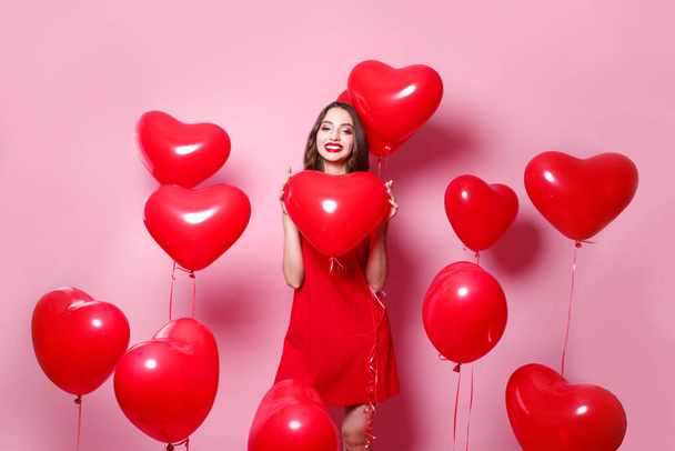 Beautiful elegant young woman in red dress with heart shape red air balloons on pink background. - Zdjęcie, obraz