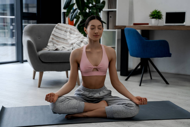 Young smiling attractive sporty woman practicing yoga, doing Ardha Padmasana exercise, meditating in Half Lotus pose working out, wearing sportswear. Healthy people lifestyle, strong woman body - Photo, Image