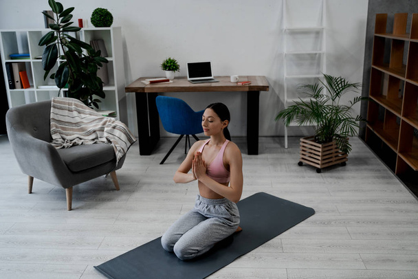 Jeune femme faisant du yoga avec le geste namaste, assis dans l'exercice de yoga, pose d'asana, formation, portant des vêtements de sport, pantalon gris, soutien-gorge de sport haut, fond intérieur à la maison
 - Photo, image
