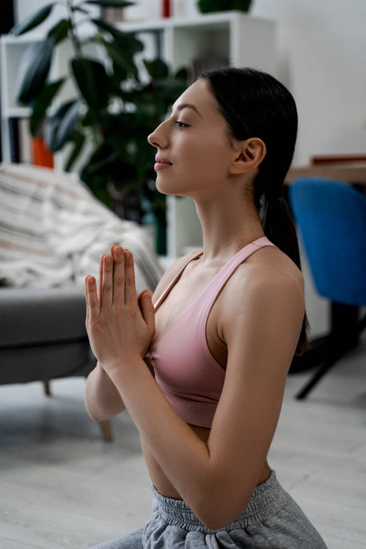 Young woman doing yoga with namaste gesture, sitting in yoga exercise, asana pose, training, wearing sportswear, gray pants, sports bra top, home interior background - Foto, afbeelding