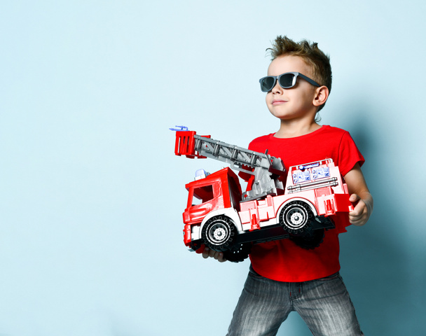 positiver Junge in heller stylischer Freizeitkleidung und Sonnenbrille stehend und mit Spielzeug-Feuerwehrauto in der Hand vor blauem Hintergrund - Foto, Bild