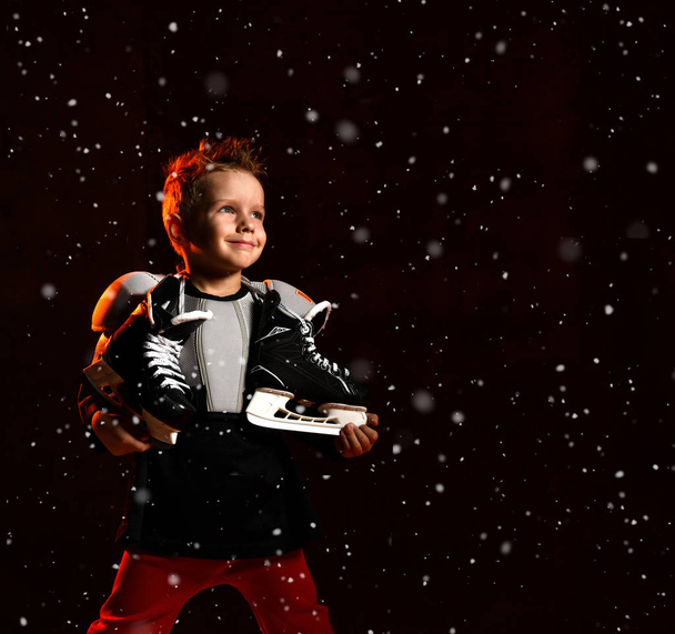 Blond jongetje in hockey uniform met hockey schaatsen op de nek staande en kijkend naar camera over donkere achtergrond - Foto, afbeelding