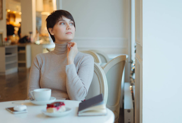 Woman wearing beige turtleneck sitting in cafe looking through window - Фото, зображення