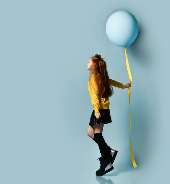 Teenage girl in yellow sweatshirt, black skirt, knee-highs, boots. She holding balloon, posing sideways on blue background. - 写真・画像