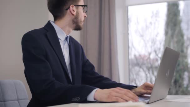 Portrait of young handsome Caucasian man looking out the window and typing on laptop keyboard. Confident businessman in suit and eyeglasses working online. Lifestyle, intelligence, wealth. - Metraje, vídeo