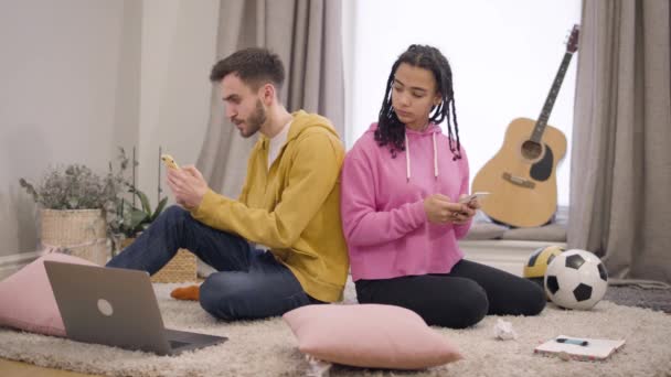 Young Caucasian man sitting back to back with girlfriend and surfing Internet. Beautiful African American girl pushing his shoulder, talking, and leaving. Relationship problems, secrets, trust. - Metraje, vídeo
