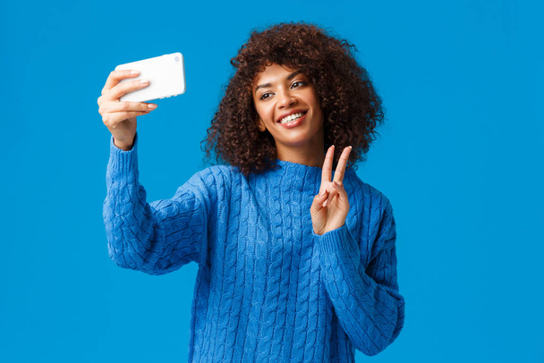 Cheerful friendly and cute african-american female student taking photo herself apply filters in new smartphone app, taking selfie tilt head lovely smiling, making peace gesture, blue background - Photo, Image