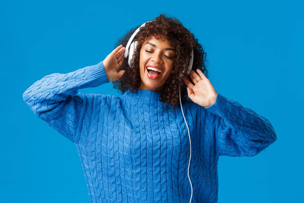 Portrait taille haute insouciante femme souriante heureuse relaxante, chantant le long, jouant application karaoké dans les écouteurs, fermer les yeux passionnément lip-sync à la musique dans les écouteurs, debout fond bleu - Photo, image