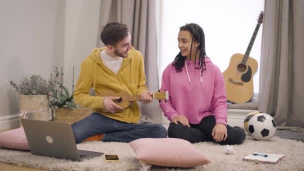 Portrait of positive multiethnic couple playing ukulele and singing at home. Smiling young African American girl and Caucasian boy having fun together on weekends. Lifestyle, music, hobby. - Metraje, vídeo