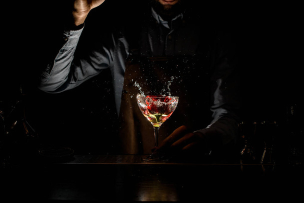 professioneller Barkeeper, der im Dunkeln eine rote Rosenknospe in ein Martini-Glas mit einem Cocktail wirft - Foto, Bild