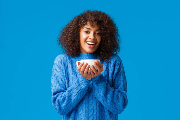 Happiness, comfort and cozy home concept. Happy relaxed and cheerful african american woman enjoying nice hot cup tea, smiling and gazing delighted, standing satisfied over blue background - Photo, Image