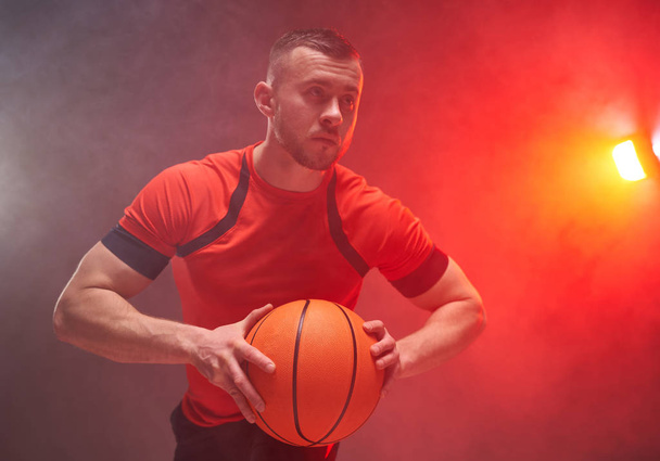 Νεαρός αθλητικός άνδρας, μπασκετμπολίστας ετοιμάζεται για ρίψη της μπάλας με πίσω φώτα και καπνό στο παρασκήνιο - Φωτογραφία, εικόνα