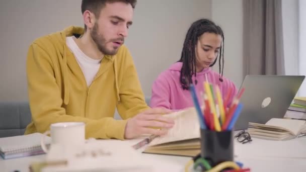 Camera moving up, two young groupmates sitting at the table and studying. Caucasian tired boy yawning and exhausted African American pretty girl with afro pigtails rubbing eyes. Tiredness, fatigue. - Кадры, видео