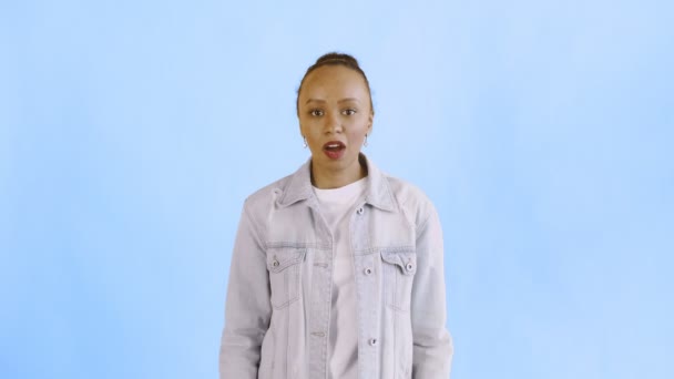 Frightened african american woman afraid of something and looks into the camera with big eyes full of horror over blue background. Jean jacket - Footage, Video