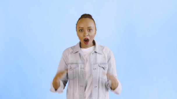 Frightened african american woman looks into the camera with open mouth and lifts hands to head on blue background. Jean jacket - Footage, Video