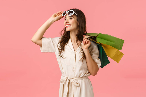 Carefree enthusiastic woman in dress, taking off glasses and looking happily sideways as enjoying beautiful day, fantastic shopping with discounts, holding shop bags behind back, pink background - Photo, Image