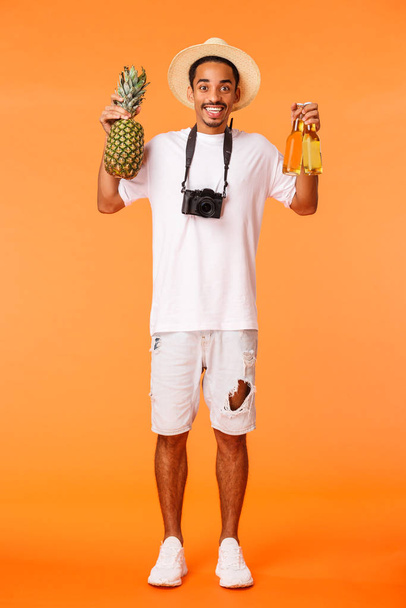 Full-length κάθετη βολή χαρούμενος αφροαμερικανός τύπος προτείνοντας ποτό και να απολαύσετε τις διακοπές, κρατώντας ανανά και δύο μπουκάλια μπύρα, χαμογελώντας ενθουσιώδης, χαλαρώνοντας στον ελεύθερο χρόνο, πορτοκαλί φόντο - Φωτογραφία, εικόνα
