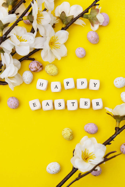 Happy Easter. Cubes with text on yellow background. Spring brunches with white flowers and candy, chocolate easter eggs. Minimalistic flat lay. - Photo, image