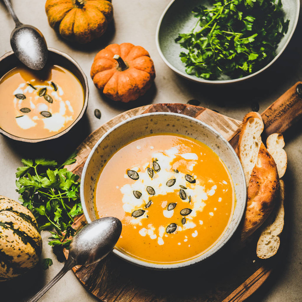 Autumn, winter warming seasonal meal. Flat-lay of pumpkin soup with seeds, parsley and cream in bowls over grey concrete background, top view, square crop. Vegetarian, healthy, comfort food concept - Foto, Imagem