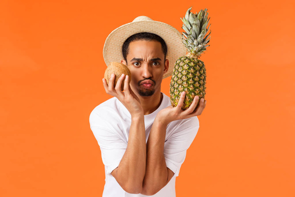 Engraçado e fofo, cara afro-americano bobo brincando, curtindo o verão viajando para o exterior, experimentando frutas tropicais como visitar o país quente, viagem de livro, segurando coco e abacaxi
 - Foto, Imagem