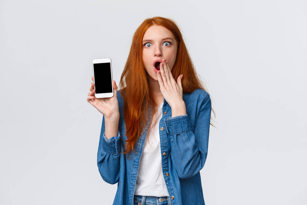 Impressed and astonished redhead woman seeing something unbelievable, spilling tea in internet, gasping cover opened mouth and showing samrtphone display, white background - Photo, Image