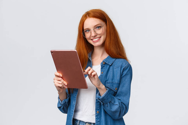 Przyjazny dla talii portret ruda uczennica przeglądająca sklepy internetowe, trzymająca tablet cyfrowy, wybierająca nowy kurs internetowy do nauki, uśmiechnięta, beztroska kamera, białe tło - Zdjęcie, obraz