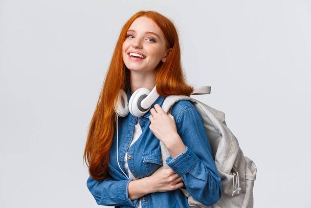 Φοιτητική ζωή, μοντέρνος τρόπος ζωής και εκπαίδευση. Χαρούμενη όμορφη κοκκινομάλλα μαθήτρια με μακριά μαλλιά, με ακουστικά στο λαιμό, σακίδιο πλάτης, χαμογελαστή κάμερα - Φωτογραφία, εικόνα