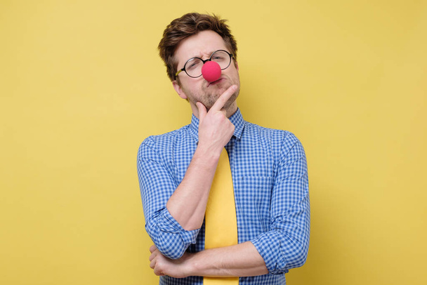 Hombre caucásico divertido en nariz de payaso rojo y corbata amarilla pensando con una mano en la barbilla
 - Foto, imagen