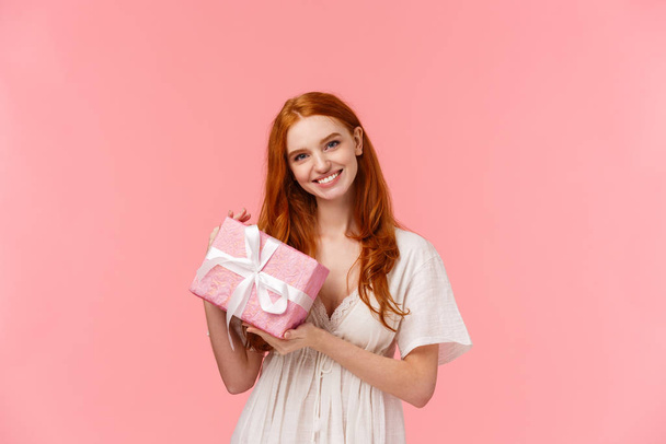 Прекрасна, ніжна і мила романтична дівчина з рудим волоссям, біла сукня, тримає подарунок, дарує партнеру подарунок і посміхається, вітає з річницею, стоїть рожевий фон
 - Фото, зображення