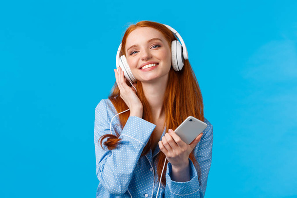 Technologie, Musik und Unterhaltungskonzept. Fröhlich attraktive rothaarige kaukasische Frau, die mit Kopfhörern Musik hört, Smartphone in der Hand, erfreut lächelnd, blauer Hintergrund stehend - Foto, Bild