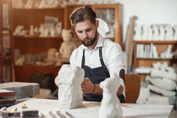 Male sculptor in white shirt and black apron makes a limestone copy of woman torso at the artistic studio. - Фото, изображение
