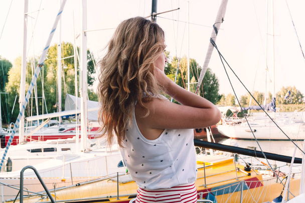back view of woman with long hair in white blouse and red skirt standing and lookig away in front of sailboats in yachts club near river - Φωτογραφία, εικόνα