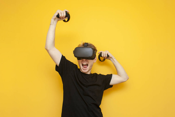 fröhlicher junger Kerl in vr Brille auf gelbem Hintergrund spielt und gewinnt mit Joysticks, ein emotionaler Mann gewinnt ein virtuelles Spiel - Foto, Bild