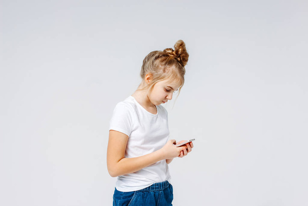 Carefree smiling preteen girl with fair hair in white t-shirt and jeans using mobile isolated on white background - Fotoğraf, Görsel
