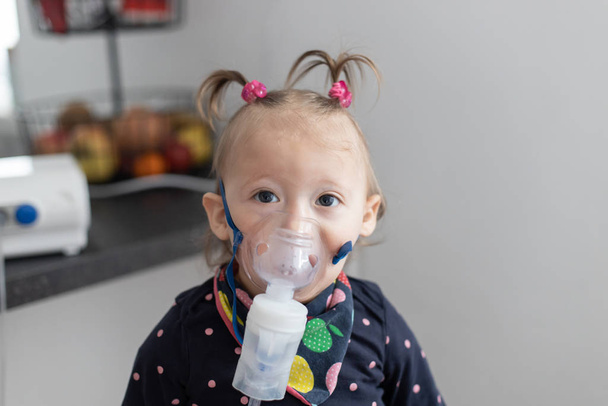 Cute little baby in the mask of an inhaler. Procedure of inhalation at home. Baby taking respiratory therapy with nebulizer.  - Photo, image