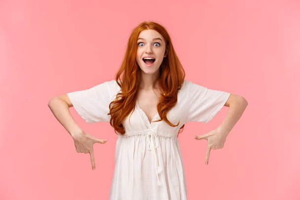 Excited and happy, amazed cheerful redhead woman seing her favorite product in store, pointing fingers down looking with temptation and desire, eager buy it, pink background - Zdjęcie, obraz