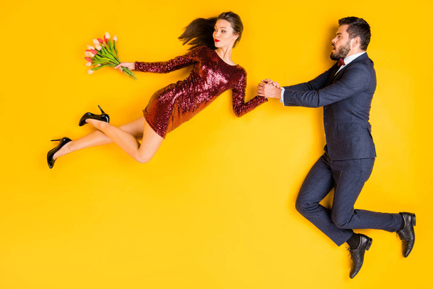 Top above high angle profile side full body photo of two people date afraid man hold hand catch cunning woman fly wind carry tulips bunch wear tuxedo dress lay isolated yellow color background - Valokuva, kuva