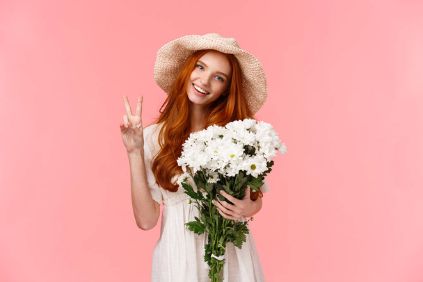 Voorjaar, feest en emoties concept. Vrolijke mooie roodharige vrouw in schattig stro hoed, genieten van romantische date, met boeket witte bloemen, toon vrede teken en glimlach gelukkig, roze achtergrond - Foto, afbeelding